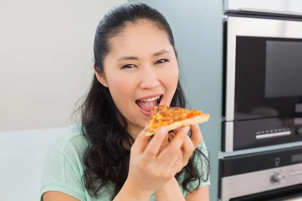 Mujer joven sonriente comiendo una rebanada de pizza en la cocina —  Fotos de Stock