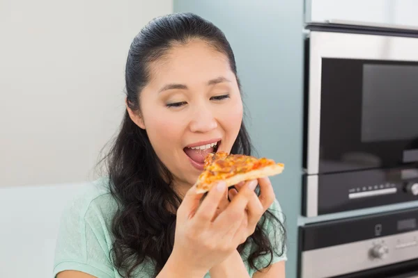Mujer joven sonriente comiendo una rebanada de pizza en la cocina — Foto de Stock