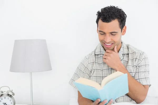 ベッドで本を読んでリラックスした男の笑みを浮かべてください。 — ストック写真
