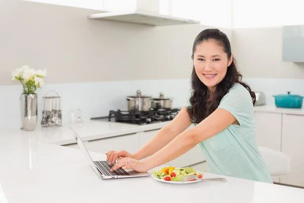 Mutfakta salatası yerken dizüstü kullanan kadın — Stok fotoğraf