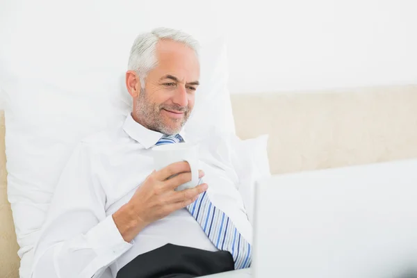 Smiling mature businessman using laptop in bed — Stock Photo, Image