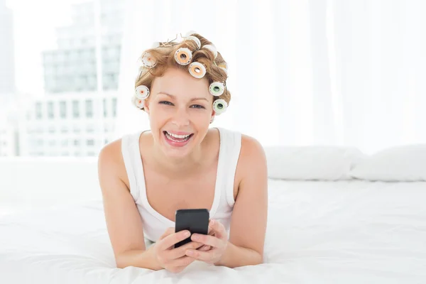 Cheerful woman in hair curlers text messaging in bed — Stock Photo, Image