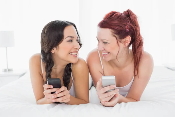 Two smiling female friends text messaging on bed — Stock Photo, Image