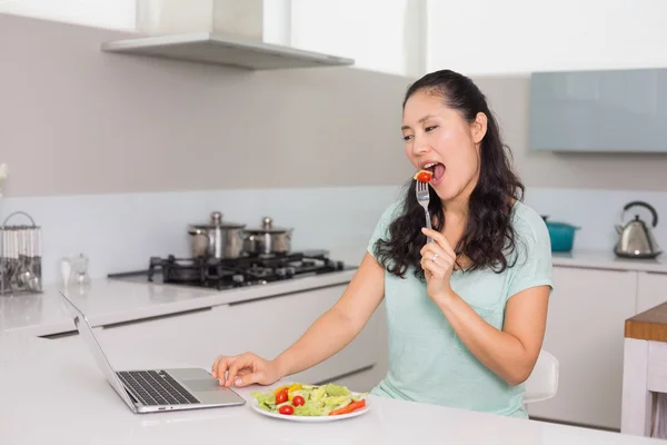 Ung kvinna med laptop äta sallad i köket — Stockfoto