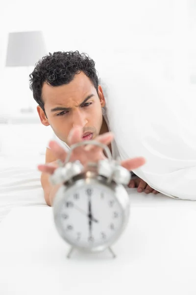Hombre somnoliento en la cama extendiendo la mano al reloj despertador Imágenes de stock libres de derechos