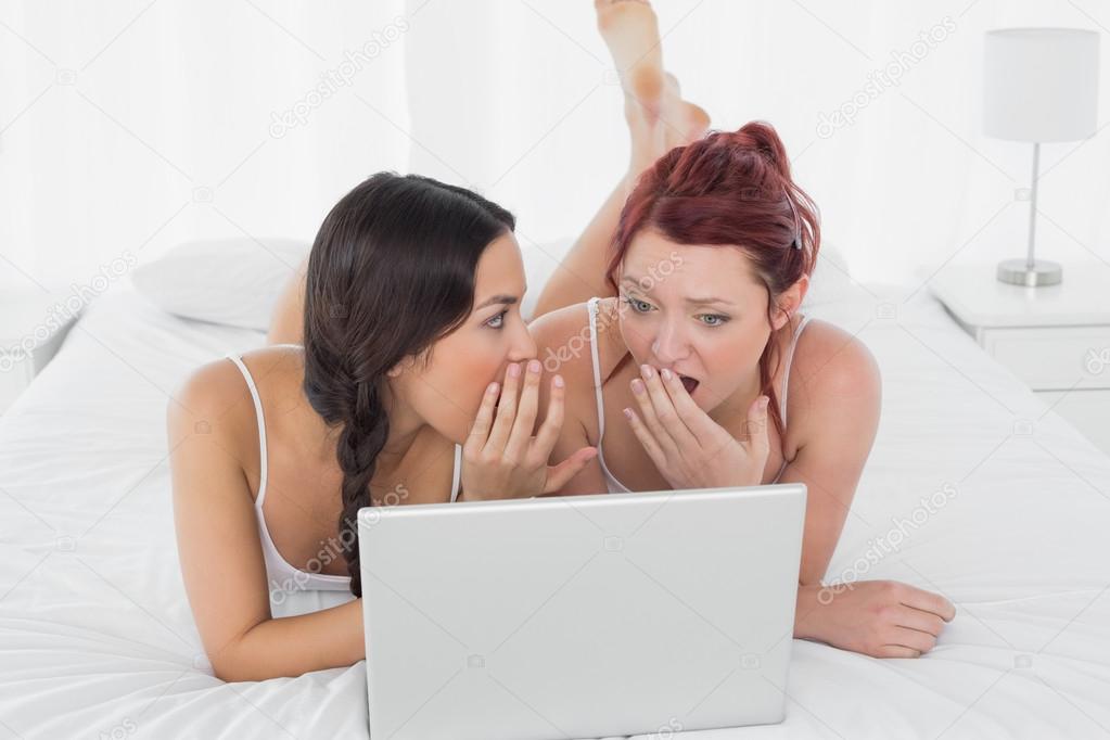 relaxed female friends with laptop gossiping in bed
