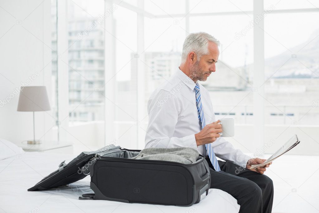 Businessman with coffee cup reading newspaper by luggage at hote