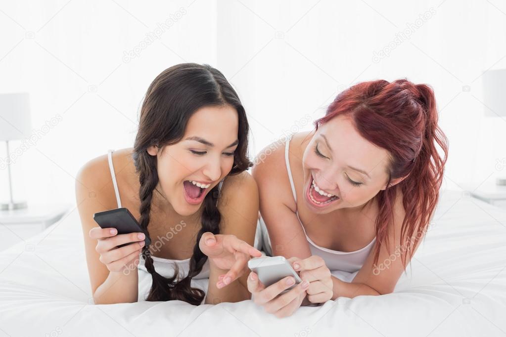 Cheerful young female friends text messaging on bed