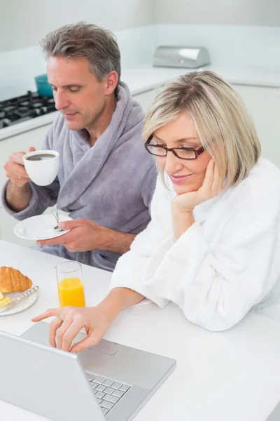 Paar in Bademänteln mit Kaffee und Saft mit Laptop in der Küche — Stockfoto