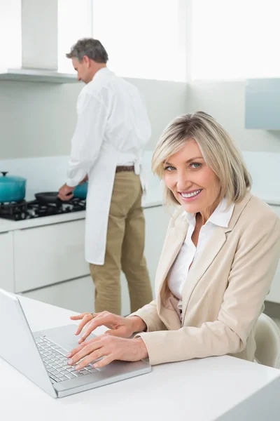 Woman using laptop and man cooking food in kitchen — Stock Photo, Image