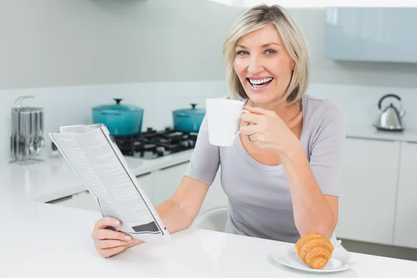 Happy woman with coffee cup and newspaper in kitchen — Stock Photo, Image