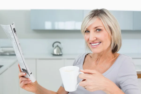 Mujer sonriente con taza de café y periódico en la cocina —  Fotos de Stock