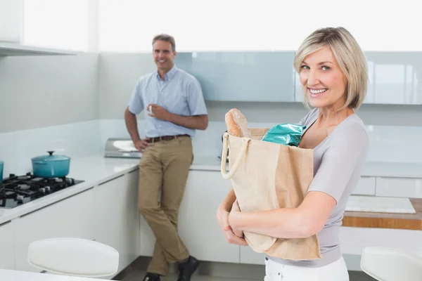 Mujer sonriente con bolsa de comestibles y hombre de fondo en la cocina — Foto de Stock