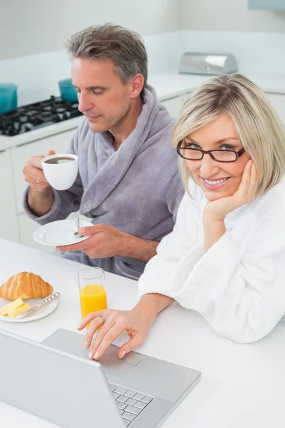 Пара в халатах с кофе и соком с помощью ноутбука на кухне — стоковое фото