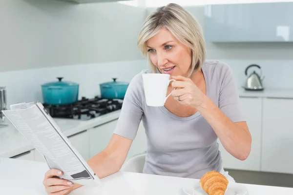 Kvinna dricker kaffe medan du läser tidningen i köket — Stockfoto