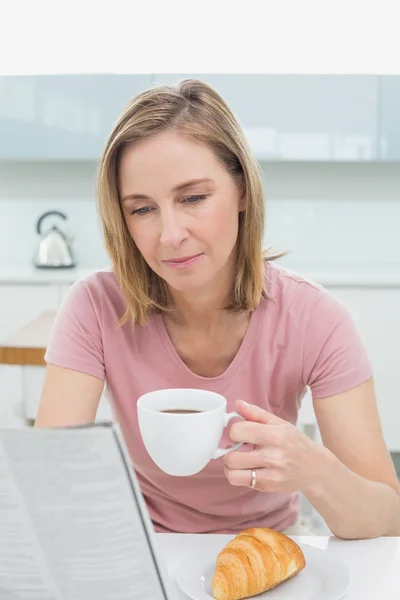 Mulher lendo jornal enquanto toma café na cozinha — Fotografia de Stock