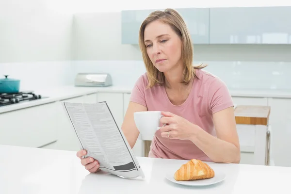 Mujer concentrada leyendo el periódico mientras toma café en la cocina —  Fotos de Stock
