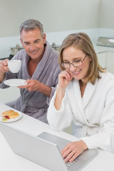 台所でラップトップを使用しながら朝食を持っているカップル — ストック写真