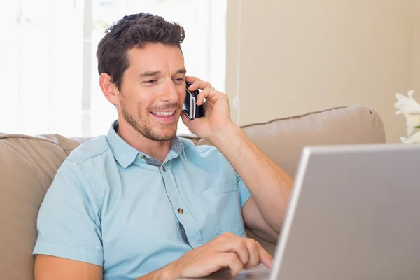 Homem feliz usando laptop e telefone celular no sofá — Fotografia de Stock