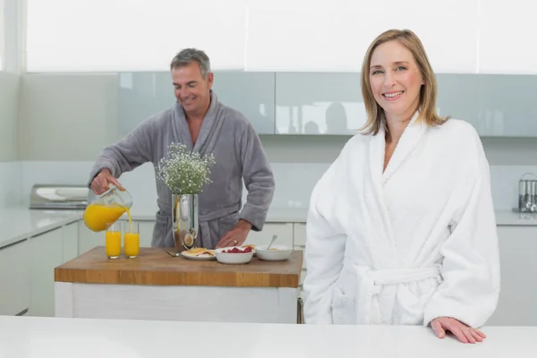 Mulher sorridente com homem derramando suco de laranja no fundo na cozinha — Fotografia de Stock