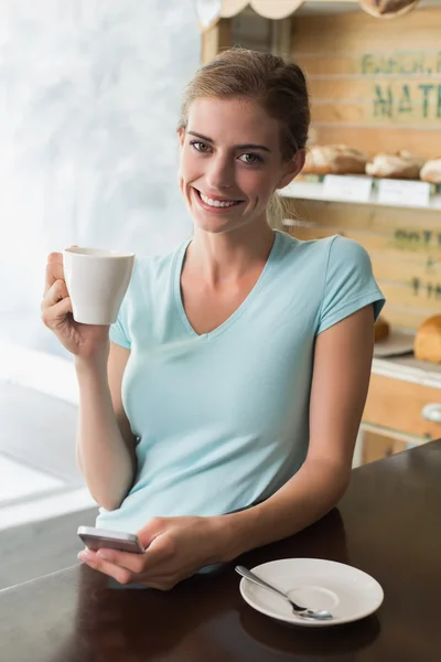 Mujer bebiendo café mientras usa el teléfono móvil en el mostrador en la cafetería —  Fotos de Stock