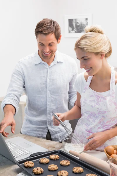 Casal preparando biscoitos ao usar laptop na cozinha — Fotografia de Stock