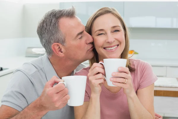 Мужчина целует счастливую женщину, когда пьет кофе на кухне — стоковое фото