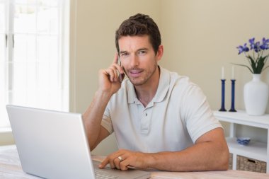 Smiling man using laptop and mobile phone at home clipart