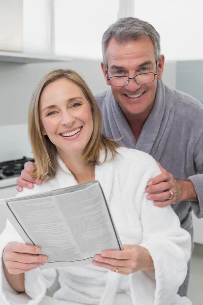 Happy couple in bathrobes reading newspaper in kitchen — Stock Photo, Image