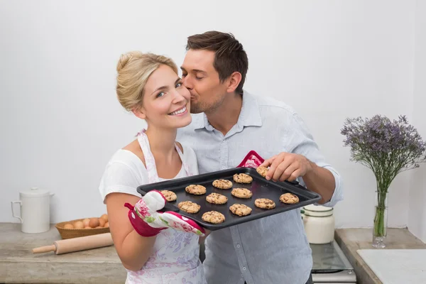 Man kissing woman's cheek as she holds freshly baked cookies in — Stock Photo, Image