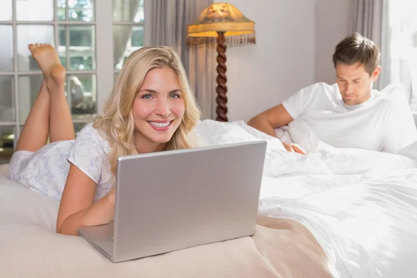 Woman using laptop while man reading book in bed — Stock Photo, Image