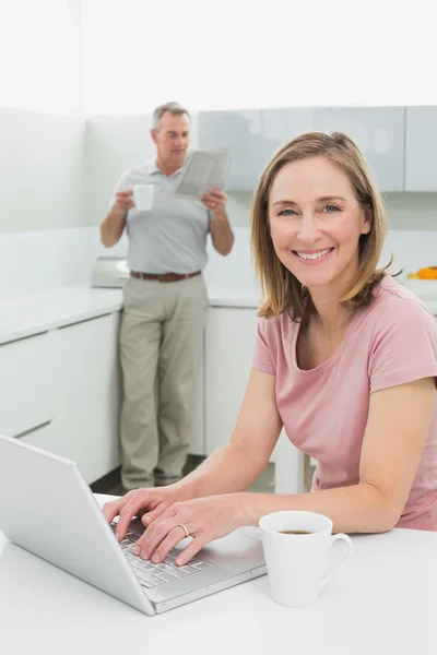 Žena pomocí notebooku, zatímco muž s šálek kávy a noviny v kuchyni — Stock fotografie