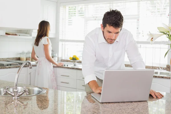 Homem usando laptop com mulher no fundo na cozinha — Fotografia de Stock