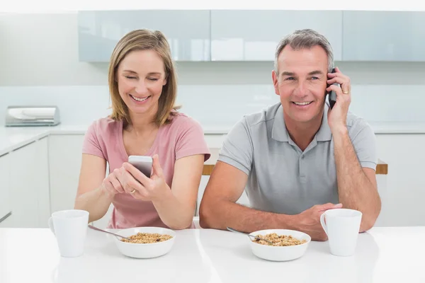 Пара, использующая мобильные телефоны во время завтрака на кухне — стоковое фото