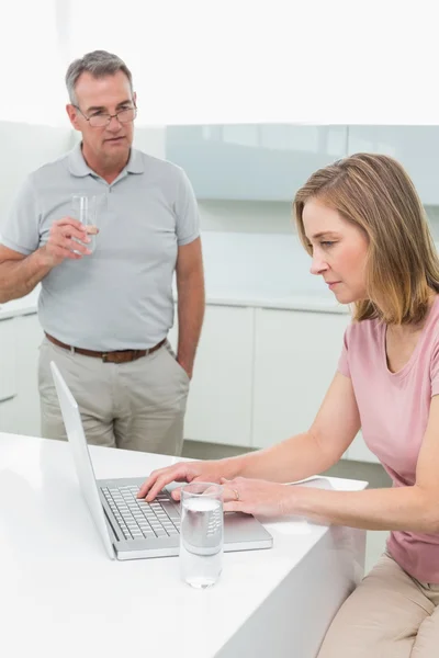 Woman using laptop and man drinking water in kitchen — Stock Photo, Image