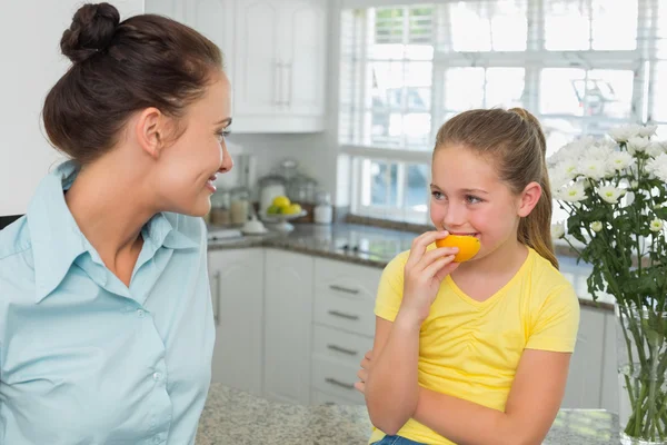 Matka, patrząc na córkę jedzenie pomarańczy w kuchni — Zdjęcie stockowe