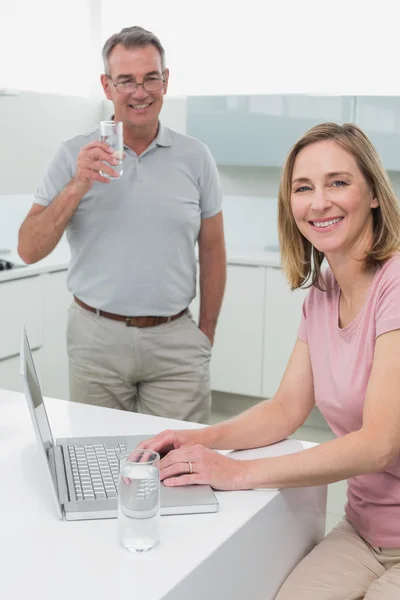 Woman using laptop while man drinking water in kitchen — Stock Photo, Image