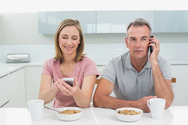 Coppia utilizzando i telefoni cellulari mentre fa colazione in cucina — Foto Stock