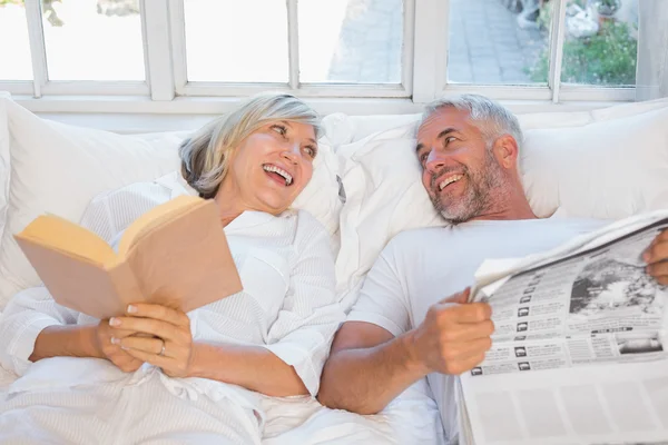 Cheerful mature couple with newspaper and book in bed — Stock Photo, Image