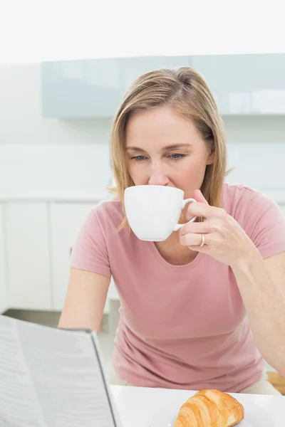 Mujer leyendo el periódico mientras toma café en la cocina —  Fotos de Stock