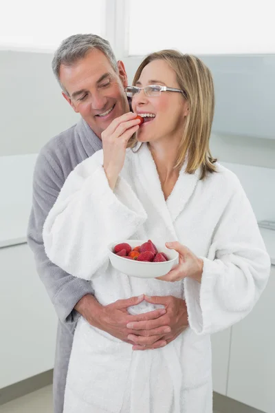 Uomo abbracciando donna come lei mangia fragola in cucina — Foto Stock