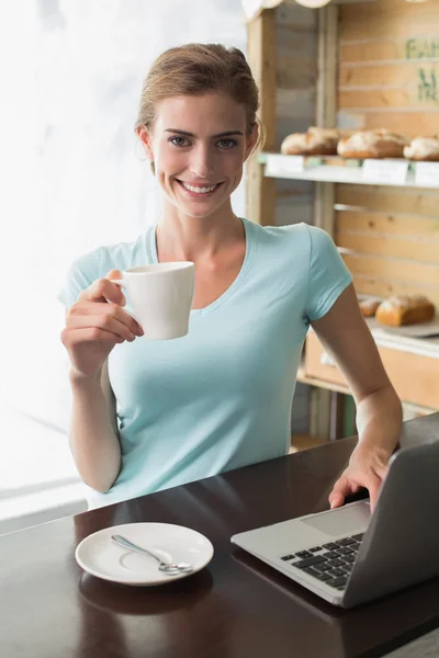 Mulher sorridente com xícara de café usando laptop na cafeteria — Fotografia de Stock