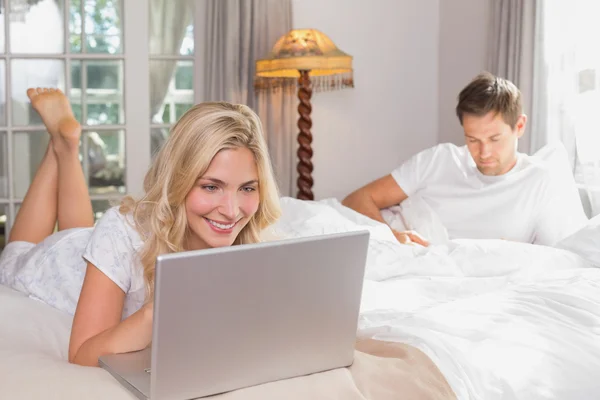 Woman using laptop while man reading book in bed — Stock Photo, Image