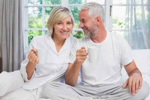 Smiling mature couple with coffee cups sitting on bed — Stock Photo, Image