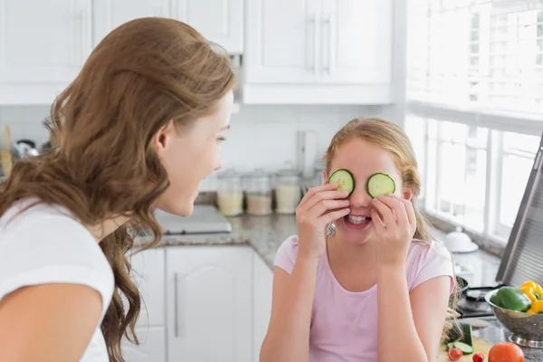 Woman looking at girl keep cucumber slices on eyes in kitchen — Stock Photo, Image