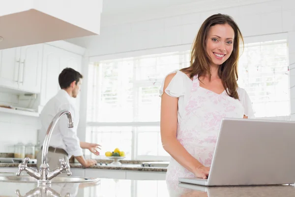 Kobieta za pomocą laptopa z mężczyzną w tle w kuchni — Zdjęcie stockowe