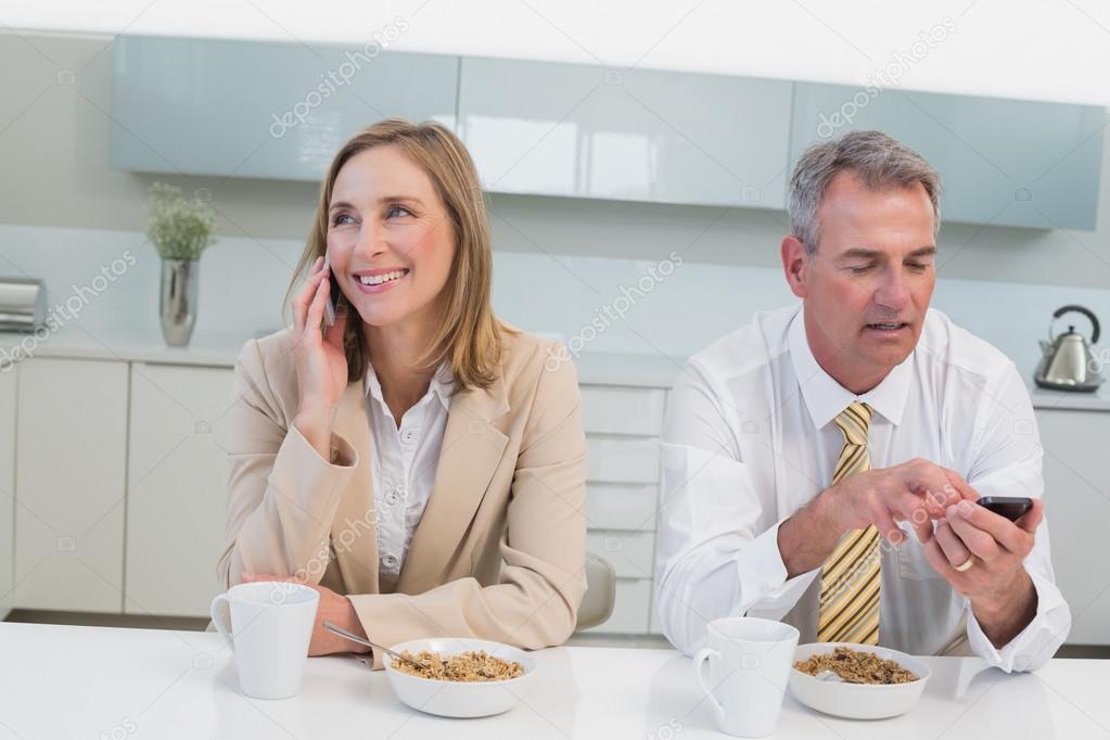 Business couple using cellphones while having breakfast in kitchen