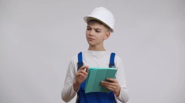 Sert şapkalı ve tulumlu, tabletle düşünen ve kameraya bakan ciddi bir çocuk. Beyaz arka planda, müteahhit üniforması içindeki özgüvenli, düşünceli beyaz çocuk portresi. Foreman işi.