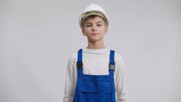 Vit pojke i hård hatt och overaller sätta händerna på höfter poserar på vit bakgrund. Medium skott porträtt av självsäker söt unge väljer byggnad yrke tittar på kameran leende. — Stockvideo