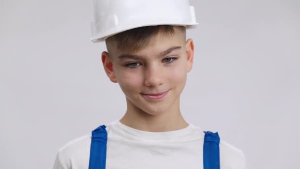 Närbild porträtt av stilig vit pojke i hård hatt poserar på vit bakgrund. Glad söt unge i byggaruniform tittar på kameran leende. Yrkesval och beslutsfattande. — Stockvideo